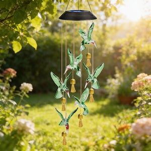 May include: A solar-powered wind chime featuring six green hummingbird ornaments with red beaks and gold tassels. The wind chime hangs from a black disc, creating a decorative outdoor accent. The background shows a sunny garden setting.