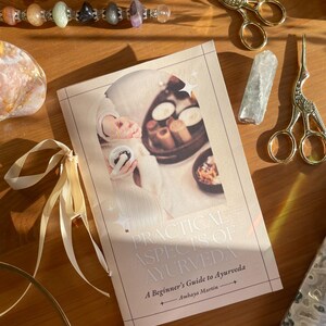 May include: A book titled "Practical Aspects of Ayurveda" with a beige cover, featuring an image of a person holding food. Gold scissors, a crystal, and a beaded bracelet are arranged around the book on a wooden surface.