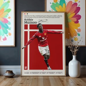 May include: Framed poster of Kobbie Mainoo in a red football jersey, number 37. The poster has a red background with white lines. Text includes his name, signature, and achievements. The poster is in a wooden frame.