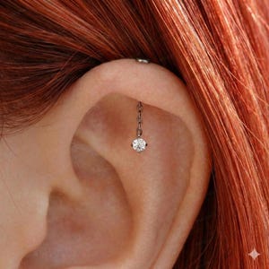 May include: A close-up of an ear with a silver chain earring. The earring features a small, sparkling, round, clear gemstone at the end of the chain. The ear has a helix piercing with a small silver stud.