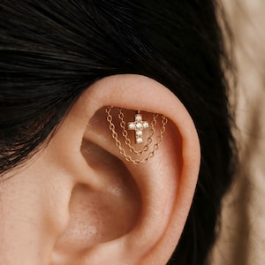 May include: A gold-toned cartilage earring featuring a small cross pendant encrusted with clear stones. The cross is suspended from two delicate gold chains that drape in a curved pattern, connecting to a single post inserted into the ear.