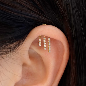May include: Gold-toned ear jewelry featuring three pieces with dangling, clear, round-cut gemstones. The earrings are designed to be worn on the helix and tragus of the ear. The jewelry is shown against a background of dark hair and skin.