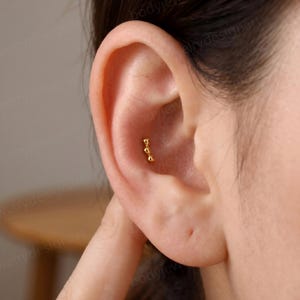 Könnte beinhalten: Ein goldfarbener Knorpel-Ohrstecker mit einer vertikalen Reihe aus drei kleinen, polierten, abgerundeten Kügelchen. Das Schmuckstück befindet sich in der Ohrmuschel und zeichnet sich durch ein schlichtes Design mit glatter, reflektierender Oberfläche aus.