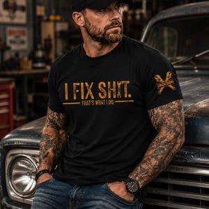 May include: A black short-sleeved t-shirt featuring the yellow distressed text "I FIX SHIT. THAT'S WHAT I DO" on the chest. The left sleeve displays a yellow graphic of two crossed wrenches with the name "JACKSON" printed between them.