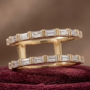 May include: A gold double-band ring featuring two parallel rows of baguette-cut clear stones. Each stone is set within a gold bar setting, creating a structured, linear design that connects the two bands with vertical gold bars on the sides.
