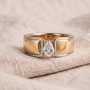 May include: A two-tone ring featuring a wide band with polished yellow gold panels framed by silver-colored metal edges. A single pear-shaped, clear faceted gemstone is set vertically in the center gap between the two gold sections of the band.