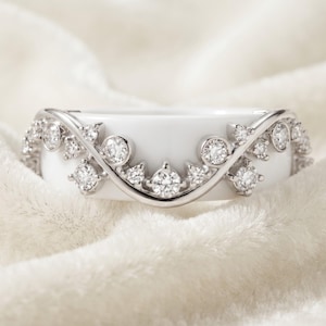 May include: A white ceramic ring featuring a silver-toned metal overlay in a curved, vine-like design. The metal structure is set with multiple small, round, clear faceted stones that follow the arching pattern across the front of the band.