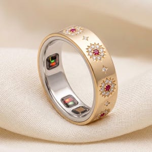 Puede incluir: Un anillo inteligente de tono dorado con una banda decorada con patrones de estrellas, cada uno centrado con una pequeña piedra roja rodeada de cristales claros. El interior del anillo muestra sensores electrónicos con luces verdes y rojas.
