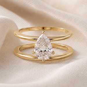 Peut inclure: Une bague en or ornée d'une pierre transparente en forme de poire au centre. L'anneau est composé de deux barres dorées parallèles qui se rejoignent au niveau du sertissage, créant un espace ouvert entre les deux bandes de chaque côté.