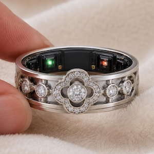 May include: A silver-toned smart ring featuring a central clover-shaped motif encrusted with small clear stones and a larger round stone in the center. The inner band displays black electronic sensor components with glowing green and red lights.