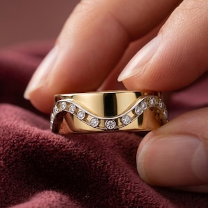 May include: A gold band ring featuring a polished finish and a central wave-shaped channel set with a row of small, round, clear white diamonds. The ring has a smooth, solid gold surface above and below the undulating diamond-encrusted wave design.