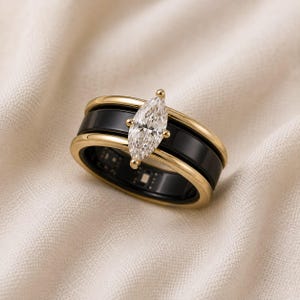 Peut inclure: Une bague avec une pierre précieuse transparente taille marquise sertie en or, entourée de deux anneaux noirs lisses. Le design présente des bords dorés en haut et en bas, avec un capteur électronique interne visible à l'intérieur de la structure.