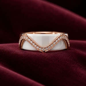 May include: A ring featuring a smooth white ceramic band accented by a rose gold-toned metal overlay in a wave pattern. The rose gold-toned metal is set with a row of small, clear, round-cut stones that follow the curved shape of the design.