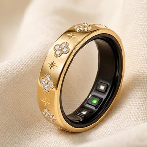 May include: A gold-toned smart ring featuring a polished exterior with engraved four-petal flower motifs set with clear stones and star-shaped accents. The interior of the ring is black with visible electronic sensors and a small green light.