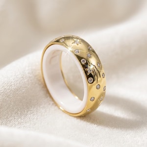 May include: A gold band ring featuring a white inner lining and a polished exterior surface decorated with multiple small, clear, round-cut stones and engraved star motifs arranged in a scattered pattern across the entire outer circumference.