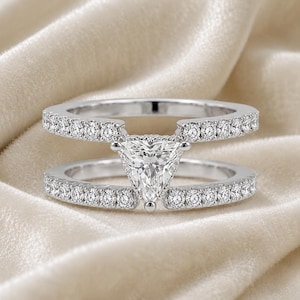 Peut inclure: Une bague argentée dotée d'un design à double anneau parallèle. Chaque anneau est orné d'une rangée de petites pierres rondes et transparentes. Une grande pierre transparente de taille triangulaire est fixée au centre, reliant les deux anneaux.