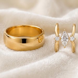 May include: A gold band ring with a smooth, polished finish alongside a gold open-style ring featuring a central marquise-cut clear stone flanked by two small round-cut clear stones on each side of the split, curved metal band design.