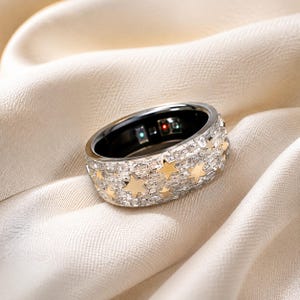 Peut inclure: Une bague large avec un extérieur argenté incrusté de nombreuses petites pierres transparentes taille baguette et plusieurs étoiles dorées incrustées. L'intérieur de la bague est noir et contient de petits composants électroniques.