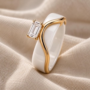 May include: A ring featuring a smooth white ceramic band accented with a polished gold-tone metal overlay in a curved, wave-like design. A single rectangular emerald-cut clear gemstone is set in a gold-tone prong setting atop the band.