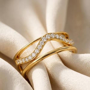 Peut inclure: Une bague dorée présentant un design multi-anneaux avec une couche supérieure ondulée incrustée de petites pierres claires taillées en rond. La bague possède une finition métallique polie et une structure superposée créant un aspect fluide.