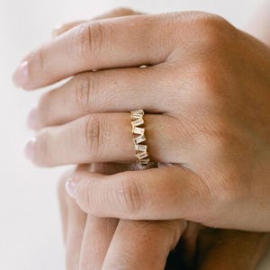 Könnte beinhalten: Ein goldener Ring mit einer Reihe rechteckiger, klarer Steine. Der Ring befindet sich an einem Finger, die Hand ist im Vordergrund. Der Hintergrund ist eine weiche, neutrale Farbe.