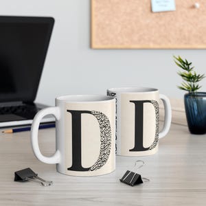 May include: Two white ceramic mugs with a large black letter "D" on a beige background. One mug features the letter filled with ornate calligraphy. The mugs are on a light wood surface with a laptop and office supplies.