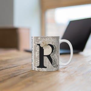 May include: White ceramic mug featuring the letter "R" in black, adorned with a floral accent and ornate calligraphy. The mug has a white handle and a decorative background with a vintage aesthetic. The mug is on a wooden surface.