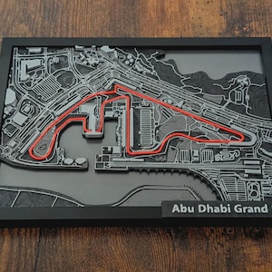 Könnte beinhalten: Eine gerahmte, dreidimensionale Karte der Rennstrecke des Abu Dhabi Grand Prix. Die Strecke ist rot auf grauem und schwarzem Hintergrund hervorgehoben, mit dem Schriftzug "Abu Dhabi Grand Prix" unten.