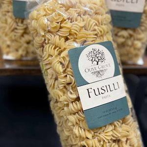 May include: Clear plastic bag filled with uncooked fusilli pasta. The bag has a teal and white label with the words "Olive Grove" and "Fusilli Pasta". The label also indicates the pasta weighs 16 oz (453.6 g).