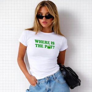 Puede incluir: Camiseta blanca con el texto verde "WHERE IS THE POT?" y un gráfico de rana verde. La camiseta se combina con vaqueros azules y un bolso negro. La persona lleva gafas de sol negras.