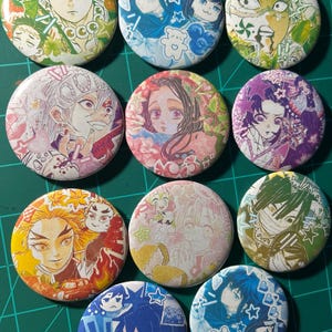 Puede incluir: Una colecci&oacute;n de pines redondos con personajes de anime coloridos. Cada pin presenta una ilustraci&oacute;n de personaje diferente con varios esquemas de color, incluyendo verde, azul, rosa y amarillo. Los pines est&aacute;n dispuestos sobre una esterilla de corte verde.