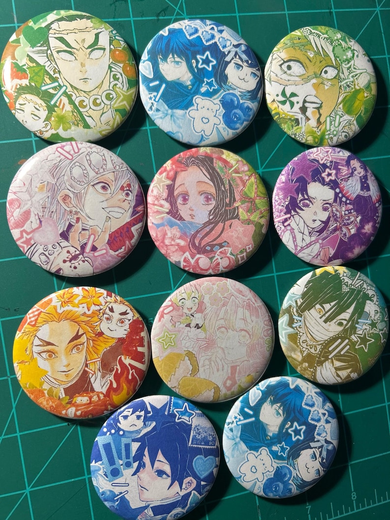 Puede incluir: Una colecci&oacute;n de pines redondos con personajes de anime coloridos. Cada pin presenta una ilustraci&oacute;n de personaje diferente con varios esquemas de color, incluyendo verde, azul, rosa y amarillo. Los pines est&aacute;n dispuestos sobre una esterilla de corte verde.