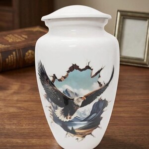 May include: White ceramic urn with a lid, featuring a detailed illustration of an eagle in flight against a mountain landscape. The design appears to break through the surface of the urn, creating a 3D effect. The urn is on a wooden surface.