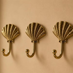 Könnte beinhalten: Drei Wandhaken aus poliertem Messing, jeder mit einer dekorativen muschelförmigen Rückplatte mit vertikalen Rillen und einem geschwungenen Haken, der in einem kleinen kugelförmigen Knopf endet, zum Aufhängen von Gegenständen.