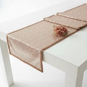 Op de afbeelding: Een roze gouden tafelloper met pailletten, een bruine rand en een klein boeket roze rozen. De rechthoekige loper ligt op een witte tafel, ideaal voor bruiloften of speciale evenementen.