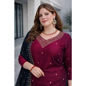 May include: A burgundy salwar kameez ensemble with a V-neckline, embroidered detailing, and small gold accents. The outfit includes a matching dark blue dupatta with gold trim. The model is wearing a necklace, earrings, and a bracelet.