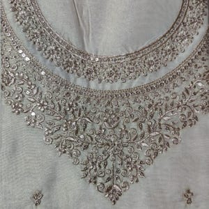 May include: A light gray fabric garment featuring an intricate neckline embellished with metallic silver embroidery and small reflective sequins. The embroidery forms a dense, symmetrical floral and vine pattern that extends downward in a V-shape.