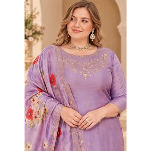 May include: A lavender-colored kurta set featuring a round neckline with intricate gold embroidery. The fabric displays a subtle sheen and is paired with a matching dupatta adorned with a vibrant floral print of red poppies and white blossoms.