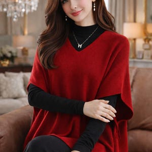 Puede incluir: Un suéter rojo estilo poncho con cuello en V sobre una camiseta negra de manga larga. El atuendo se combina con pantalones negros. Se ve un collar plateado con un colgante "H".