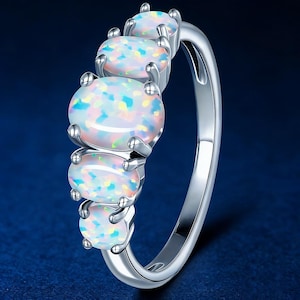 Könnte beinhalten: Ein silberner Ring mit fünf ovalen Opalen. Die Opale zeigen schillernde Farben von Blau, Grün und Gelb. Das Ringband ist aus poliertem Silber, und die Opale sind mit Krappen gefasst. Ein Schmuckstück.