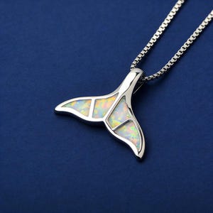 Puede incluir: Un collar con colgante plateado con diseño de cola de ballena. La cola está incrustada con piedras iridiscentes tipo ópalo, creando un efecto brillante. El collar cuelga de una delicada cadena sobre un fondo azul oscuro.