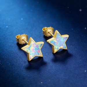 May include: A pair of gold-colored star-shaped stud earrings. Each earring features a colorful, iridescent opal center. The earrings are set against a dark blue background, creating a striking contrast. The earrings are a celestial-themed jewelry item.