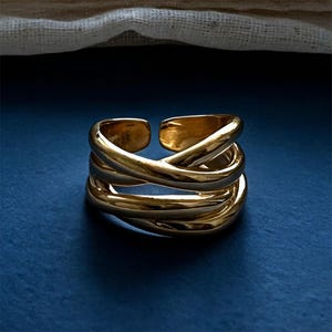 May include: A gold-colored ring with a multi-layered, crisscross design. The ring has an open design, allowing for slight size adjustments. The ring is set against a dark blue background, with a white fabric in the background.