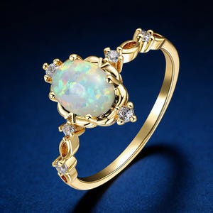 May include: A gold ring featuring an oval opal gemstone with iridescent colors. The ring is accented with small, clear gemstones and decorative gold detailing. The band has a delicate design, and the ring is set against a dark blue background.
