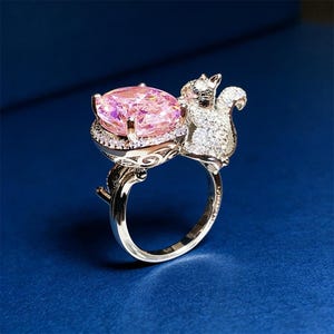 Puede incluir: Un anillo plateado con una gran gema rosa. La gema está sujeta por un engaste detallado, con un diseño de ardilla brillante en el lateral. El anillo está sobre un fondo azul oscuro.