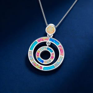 Puede incluir: Collar de plata con un colgante con círculos concéntricos con incrustaciones de piedras de ópalo de colores. El colgante tiene una piedra de ópalo circular en la parte superior y cuelga de una delicada cadena de plata. Las piedras de ópalo muestran tonos de rosa, azul y verde.