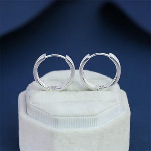 Könnte beinhalten: Ein Paar silberne Creolen. Die Ohrringe sind rund und haben ein schlichtes, poliertes Design. Sie werden auf einer weißen Samtoberfläche vor einem dunkelblauen Hintergrund präsentiert. Die Ohrringe sind ein klassisches Schmuckstück.