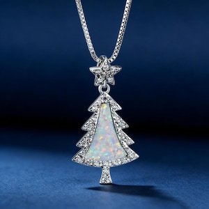 May include: Silver-toned necklace with a Christmas tree pendant. The pendant features a white opal center and is outlined with clear crystals. A star-shaped charm with crystals sits above the tree. The necklace hangs on a silver chain.