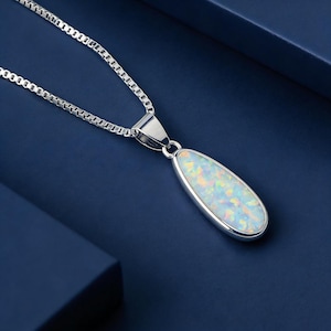 May include: A silver necklace with a pear-shaped pendant. The pendant has a white opal with iridescent colours. The chain is a box chain style. The necklace is displayed on a dark blue surface.