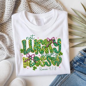 以下が含まれることがあります： 白地のTシャツに「not lucky simply blessed」の文字が緑、黄色、ピンクでプリントされています。デザインにはクローバーとリボンが含まれています。「Roman 4:7-8」の文字が下に印刷されています。Tシャツは白い面に折りたたまれています。